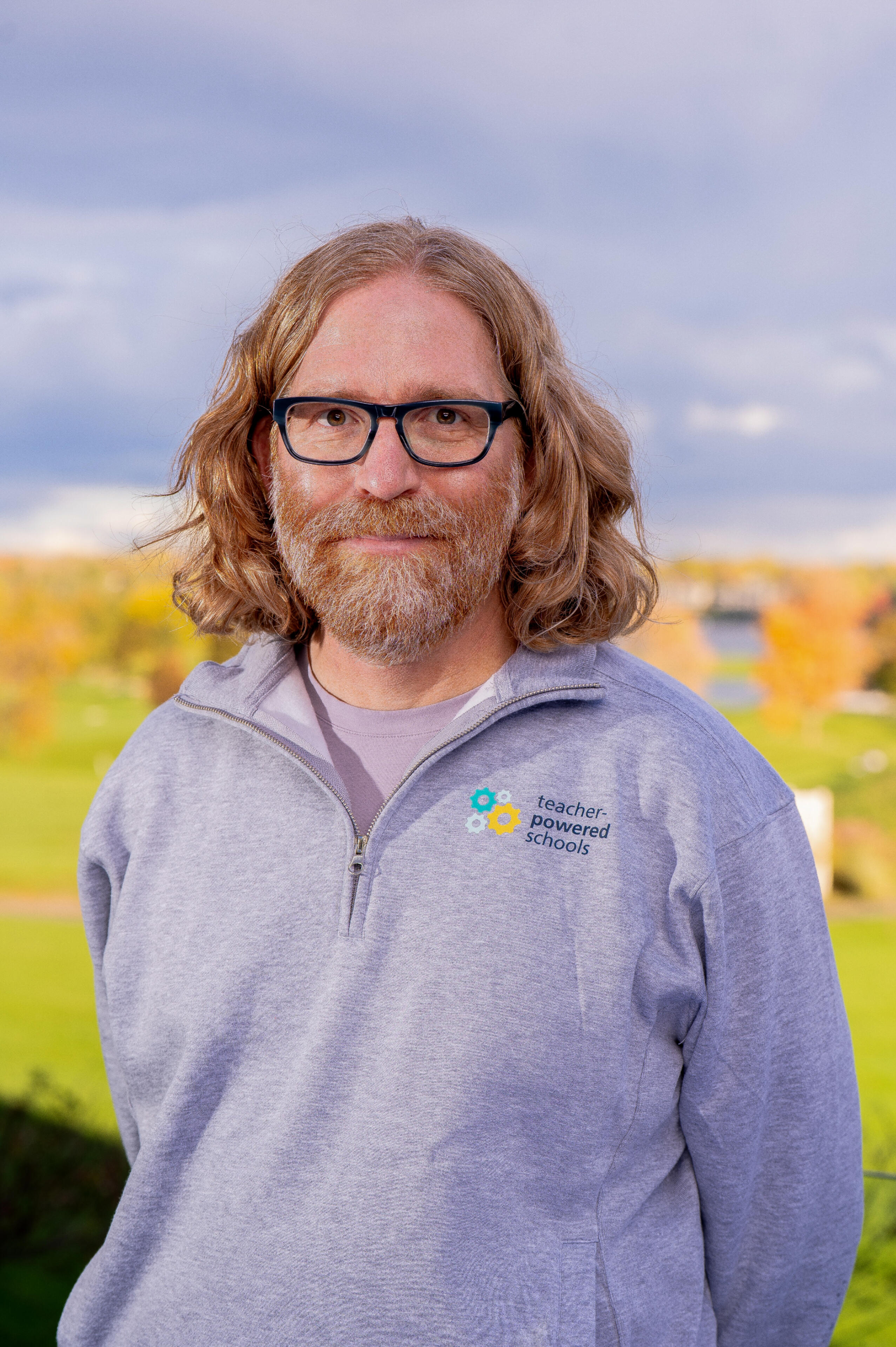 A person with long, wavy light-brown hair and a beard stands outdoors in front of a blurred autumn landscape. They are wearing glasses and a light gray quarter-zip sweatshirt with a āteacher-powered schoolsā logo on it. The lighting is warm and natural, wi