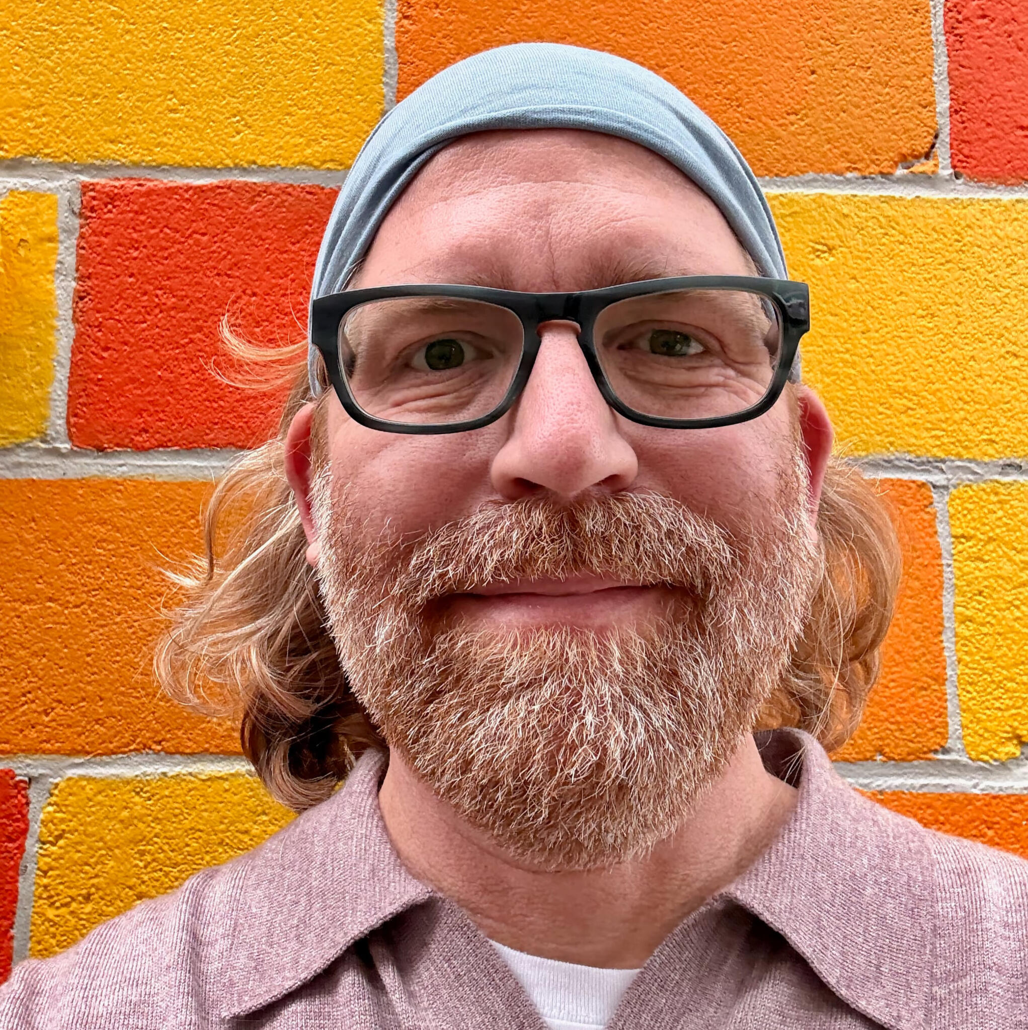 A person with long, wavy, light red hair and a light beard wearing a pale blue headband and mauve collared shirt, standing in front of a colorful brick wall painted in shades of orange and yellow.